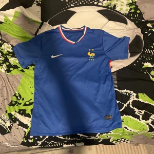 Frankrike Nike blå fotbollströja - Blå officiell Frankrike fotbollströja från Nike med broderad tupp och två stjärnor på bröstet. Tröjan har korta ärmar, röda detaljer på sidorna och en V-ringad krage med franska flaggans färger. Tillverkad i lätt och ventilerande material.