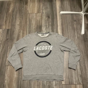 Grå sweatshirt från Lacoste Sport - Grå sweatshirt från Lacoste Sport med stort tryck på bröstet i vitt och mörkblått. Klassisk rund halsringning, långa ärmar och ribbade muddar. Mjuk insida och sportig vibe, perfekt för chill dagar. Materialet är bomullsmix. Liten fläck på O:et men annars riktigt bra tröja 