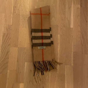 Beige rutig Burberry halsduk i kashmir - Snygg beige halsduk från Burberry i 100% kashmir, tillverkad i Skottland. Klassisk rutig design med inslag av mörkbruna, vita och orangea linjer. Mjukt material och fransar i ändarna ger en lyxig vibe. Perfekt för att höja din stil under kyliga dagar.