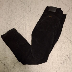 Svarta manchesterbyxor från Nudie Jeans - Svarta manchesterbyxor från Nudie Jeans i klassisk rak modell. Byxorna är tillverkade i mjuk bomull med tydlig manchesterstruktur och har snygga detaljer som logotyp på bakfickan. Perfekta för dig som gillar stilrena och bekväma byxor.