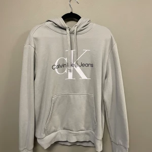 Ljusgrå hoodie från Calvin Klein - Snygg ljusgrå hoodie från Calvin Klein Jeans med stor CK-logga på bröstet. Tröjan har huva med snörning, magficka och ribbade muddar. Mjuk insida i fleece som gör den extra skön. Perfekt för chill dagar och streetstyle.