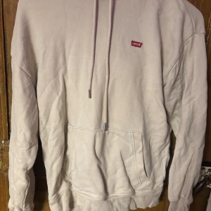 Beige hoodie från Levi's XS - Snygg ljusrosa hoodie från Levi's i storlek XS. Tröjan har klassisk huva med snörning, magficka och liten röd Levi's-logga på bröstet. Tillverkad i mjuk bomull, perfekt för chill dagar. Långärmad och loose passform för skön stil.