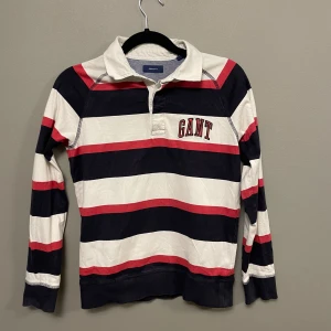 Randig pikétröja från GANT - Snygg pikétröja från GANT med breda marinblå, vita och röda ränder. Tröjan har krage med tryck på baksidan, lång ärm och GANT-brodyr på bröstet. Tillverkad i mjuk bomull, perfekt för dig som gillar klassisk och sportig stil.