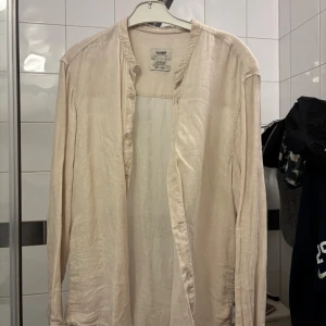 Beige linne Henley från Pull&Bear - Snygg beige Henley-skjorta från Pull&Bear i 100% linne. Skjortan har lång ärm, knappar framtill och en avslappnad passform. Perfekt för dig som gillar stilrena och luftiga plagg. Färgen är ljus och neutral, vilket gör den enkel att matcha med andra kläder.