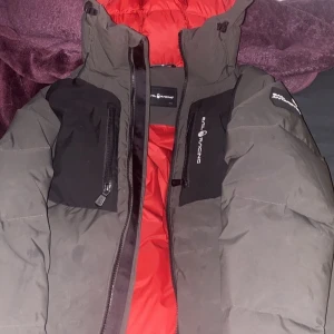 Grå pufferjacka från Sail Racing - Säljer en grå pufferjacka från Sail Racing med svart detaljpanel på bröstet och ärmen. Jackan har röd insida, huva och två bröstfickor med dragkedja. Perfekt för kalla dagar och har en riktigt schysst streetstil.