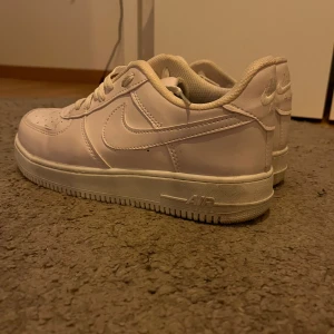 Nike Air Force 1 vita sneakers - Klassiska Nike Air Force 1 sneakers i helvitt skinn med lågt skaft och snörning. Snygg och tidlös modell med perforerad tå och tydlig swoosh-logga på sidorna. Perfekta för dig som vill ha en clean och stilren look.