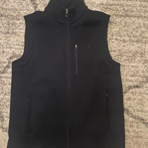 Svart fleeceväst från Polo Ralph Lauren - Svart fleeceväst från Polo Ralph Lauren Performance med hög krage och hel dragkedja framtill. Västen har tre fickor med dragkedja och en stilren, enkel design utan mönster. Perfekt för lager-på-lager och chill stil.