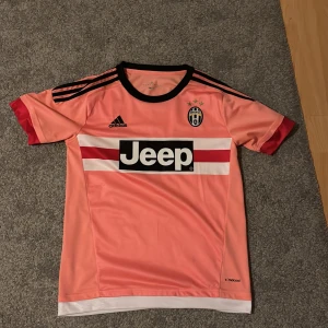 Juventus Dybala 21 rosa fotbollströja - Juventus fotbollströja med Dybala 21 på ryggen, tillverkad av Adidas. Tröjan är rosa med svarta och röda detaljer, svart krage och vita/röda ränder över bröstet. Jeep-logga på framsidan och Juventus-emblem med stjärnor. Materialet är lätt och andas, perfekt för träning.