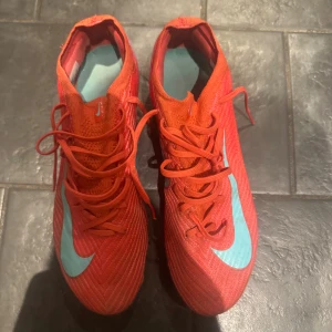 Nike Mercurial röda fotbollsskor - Säljer ett par Nike Mercurial fotbollsskor i en intensiv röd färg med ljusblå detaljer och snörning. Skorna har en spetsig tå och platt sula med dobbar för gräsplan. Ovandelen är tillverkad i syntetmaterial för lätthet och bra passform. Perfekt för dig som vill sticka ut på planen. Påse medföljer 