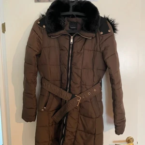 Brun quiltad dunjacka från Tindra - Snygg brun dunjacka från Tindra med svart fuskpäls på huvan, quiltad design och bälte i midjan. Jackan har långa ärmar med svarta detaljer på armbågarna och dragkedjor vid ärmslut. Perfekt för kalla dagar och har praktiska fickor framtill.