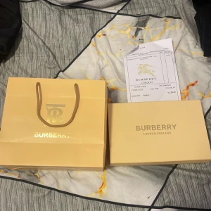 Burberry rutig skärp med B-spänne - Exklusivt Burberry bälte med klassiskt rutigt mönster i beige, svart, vit och röd. Bältet har ett stilrent metallspänne med Burberrys ikoniska B-logga i guld. Materialet är lyxigt skinn och designen är typiskt Burberry, perfekt för dig som gillar statement-accessoarer.