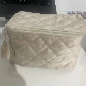 Beige quiltad necessär med tofs - Snygg beige necessär med quiltat mönster och mjuk sammetkänsla. Väskan har dragkedja med en dekorativ tofs och är perfekt för smink eller småprylar. Stilren och praktisk design som passar i väskan eller på resan.