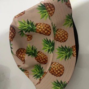 Bucket hat med ananasmönster - Snygg bucket hat i beige med färgglatt ananasmönster. Perfekt för att sticka ut på festival eller stranden. Hatten har bred brätte och är tillverkad i ett mjukt syntetmaterial som känns lätt och bekvämt på huvudet.