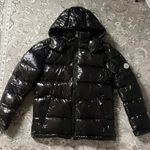 Svart Moncler Maya med huva - Säljer en svart Moncler Maya från Moncler med glansig finish och huva. Jackan har dragkedja framtill, två sidofickor med dragkedja och Moncler-logga på ärmen. Perfekt för kalla dagar och har en riktigt snygg streetstil.