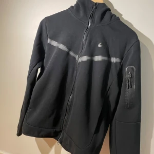 Svart Nike tech jacka  - Svart Nike tech jacka i medium med stilrena grå detaljer över bröstet och en praktisk ficka med dragkedja på armen. Jackan har hel dragkedja framtill och sportig design, perfekt för dig som gillar streetwear och funktion.