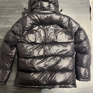 Svart pufferjacka från Polo Ralph Lauren - Snygg svart pufferjacka från Polo Ralph Lauren med röd broderad logga på bröstet. Jackan har huva, flera dragkedjeförsedda fickor och en glansig finish. Materialet är polyester och bomull, perfekt för kalla dagar. Stängs med dragkedja framtill.