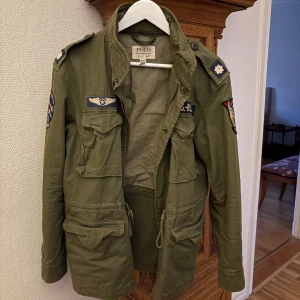 Grön filed jacket från Polo Ralph Lauren - Säljer en grön field jacka från Polo Ralph Lauren med coola patchar och detaljer på axlar och ärmar. Jackan har flera stora fickor framtill, snörning i midjan och dragkedja. Perfekt för dig som gillar militärinspirerad stil och vill sticka ut. Ny pris 6000 kr från PRL hemsida! Passar M med!