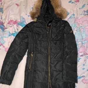 Svart lång jacka från Hollies - Svart lång pufferjacka från Hollies med huva och snygg fuskpäls. Jackan har guldfärgade dragkedjor och emblem på ärmen. Perfekt för kalla dagar med sin quiltade design och varma känsla. Fodret är i syntetmaterial och jackan har praktiska fickor med dragkedja.