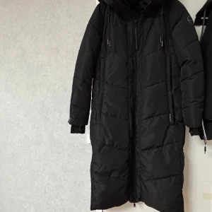 Svart lång pufferjacka från Marikoo: Nadaree - Säljer en svart lång pufferjacka från Marikoo med huva och snygga snörningsdetaljer. Jackan har dragkedja framtill, praktiska fickor med dragkedja och quiltad design. Perfekt för kalla dagar med sin rejäla längd och värmande material. Jackan är helt ny, är oanvänd. Etiketter står kvar. Är vattentät, vindtät. 