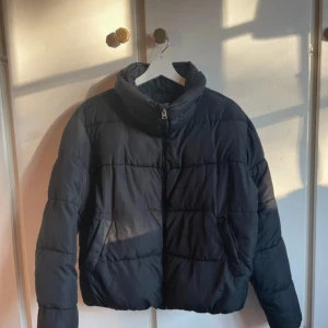 Svart pufferjacka från 157, storlek M - Svart pufferjacka från 157 i storlek M. Jackan har en hög krage, två sidofickor och dragkedja framtill. Den är quiltad och har en loose passform, perfekt för kalla dagar. Materialet är syntet och jackan är mörk i tonen. Liten defekt på sista bilden. 