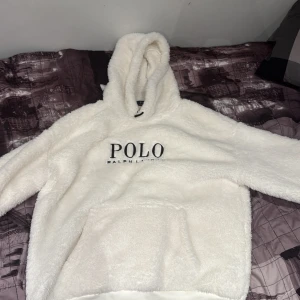 Vit fluffig hoodie från Polo Ralph Lauren - Supermjuk och fluffig vit hoodie från Polo Ralph Lauren med stor huva och broderad logga framtill. Perfekt för kalla dagar när du vill vara både bekväm och snygg. Stora fickor framtill och långärmad modell.
