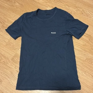 Mörkblå t-shirt från BOSS - Snygg mörkblå t-shirt från BOSS med diskret logga på bröstet. Klassisk rund hals och korta ärmar, perfekt för en clean och stilren look. Hör av er vid intresse!