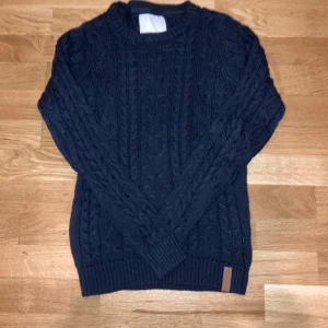 Mörkblå 157 kabelstickad tröja - Mörkblå kabelstickad tröja från 157 med klassiskt flätmönster och ribbade muddar. Tröjan har rund halsringning och en snygg läderdetalj nertill. Perfekt för dig som gillar en stilren och mysig look under kyliga dagar.
