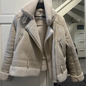 Beige teddyjacka  - Säljer en beige teddyjacka från Stradivarius i storlek S. Jackan har fluffigt teddyfoder, bred krage och detaljer med dragkedjor. Den är croppad med ribbade muddar och har snygga sömmar på ärmarna. Perfekt för dig som vill ha en varm och trendig look.