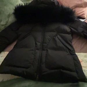 Svart pufferjacka med pälsdetalj (Monel)  - Säljer en svart pufferjacka med fluffig pälsdetalj på huvan. Jackan har dragkedja framtill och är designad för att hålla dig varm under kalla dagar. Perfekt för dig som vill ha en stilren och mysig vinterjacka med snygga detaljer.