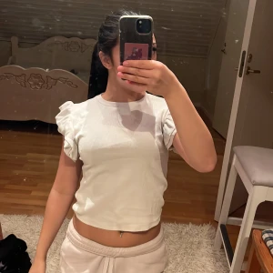 Vit ribbad topp med volangärmar Gina Tricot - Supersöt vit ribbad topp från Gina Tricot i storlek XS. Toppen har korta volangärmar och croppad passform som ger en trendig look. Perfekt att styla med jeans eller kjol för en fräsch vibe.