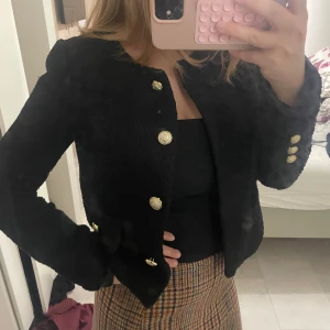 Svart cardigan jacka med guldknappar - Snygg svart cardigan jacka med strukturerat tyg och dekorativa guldknappar framtill och på ärmsluten. Jackan har en klassisk rak passform, rund halsringning och fickor framtill. Perfekt för dig som gillar stilrena detaljer och vill ha en jacka som sticker ut. S/M