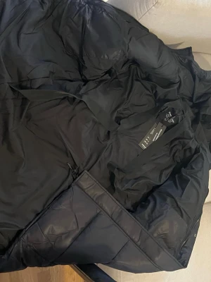 Svart lång pufferjacka  - Säljer en svart lång pufferjacka  med huva och dragkedja samt tryckknappar framtill. Jackan har två sidofickor och breda quiltade paneler som ger en riktigt puffig look. Perfekt för kalla vinterdagar och riktigt skön att bära.