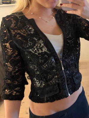 Svart spetströja med dragkedja, trekvartsärmad - Svart kofta/blus/tröja från Topshop i välgjord spets med dragkedja framtill. Blusen har trekvartsärm och dekorativa fickor framtill. Snygga satindetaljer längs kanterna ger en extra touch. Festtopp eller snygg vardags kofta.