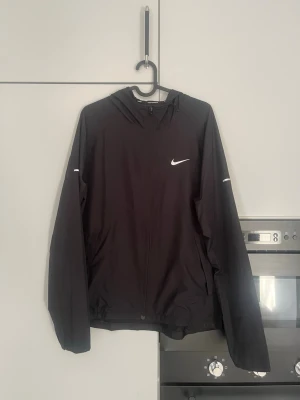 Nike Miller - Svart vindjacka från Nike med huva och dragkedja framtill. Jackan har två sidofickor och en vit Nike-logga på bröstet. Lätt material och diskreta reflexdetaljer på ärmarna. Perfekt för blåsiga dagar och sportiga outfits.