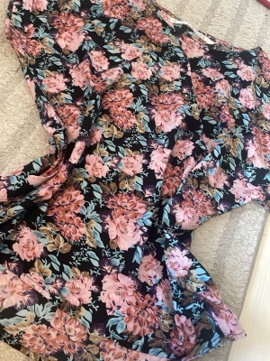 Svart blus med rosa o blå blommor - Snygg blus/t-shirt med svart botten och stora blommor i rosa och blått. Luftigt tyg och avslappnad passform, perfekt för dig som gillar färg och mönster. Blusen har korta ärmar och rund halsringning. Materialet känns lätt och flowy.