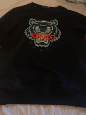 Svart Kenzo sweatshirt med tiger - Svart sweatshirt från Kenzo med ikoniskt tigerhuvud broderat på bröstet och Kenzo Paris-text i rött. Tröjan har rund halsringning och ribbade muddar vid ärmslut och nederkant. Perfekt statement-plagg för dig som gillar streetwear.