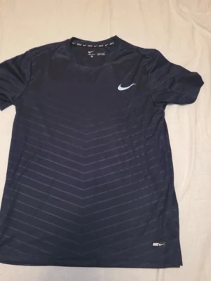 Svart Nike t-shirt med mönster - Snygg svart t-shirt från Nike med diskreta, tunna ränder i chevronmönster. T-shirten har rund hals, korta ärmar och en vit Nike-logga på bröstet och ryggen. Tillverkad i ett lätt och sportigt material som andas.