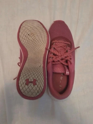 Rosa Under Armour sneakers - Säljer ett par snygga rosa sneakers från Under Armour med vit sula och logga på sidan och undersidan. Skorna har meshmaterial på ovandelen för bra andningsförmåga och klassisk snörning. Perfekta för dig som vill ha en sportig och trendig look.