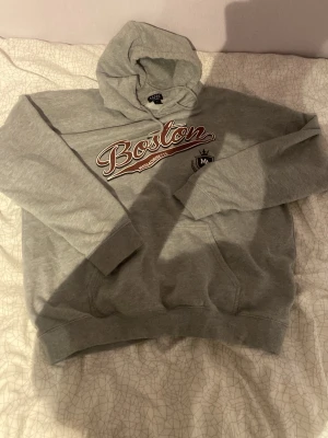 Grå Boston hoodie med tryck - Säljer en grå hoodie från Oasis med stor Boston-text och Massachusetts-tryck på bröstet. Klassisk känguruficka framtill och huva med dragsko. Perfekt för dig som gillar college-stil och vill ha en skön och avslappnad look. Grejen är att det står s men den är riktigt oversized så egentligen är den L.