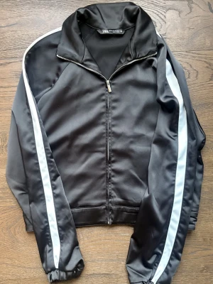 Svart träningsjacka från Zara - Svart träningsjacka från Zara med vita ränder längs ärmarna. Jackan har dragkedja framtill, hög krage och resår vid ärmsluten. Tillverkad i ett glansigt, sportigt material som passar perfekt till träning eller en sportig look.