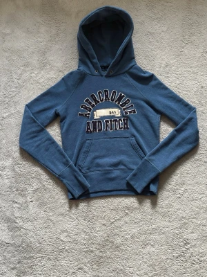 Blå hoodie från Abercrombie & Fitch - Snygg blå hoodie från Abercrombie & Fitch med stor logga och tryck framtill. Klassisk känguruficka och huva med dragsko. Mjuk bomullsmix som känns skön mot huden. Perfekt för en avslappnad och cool stil. Skulle säga även om det står medium så passar den bättre på nån som är xs/s eftersom den är väldigt tight runt midjan och lite kort