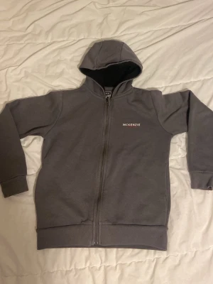 Grå zip-hoodie från McKenzie - Snygg grå hoodie med dragkedja från McKenzie. Klassisk modell med huva, ribbade muddar och diskret logga på bröstet. Perfekt för en chill och avslappnad stil. Mjuk bomullsblandning som känns skön mot huden.