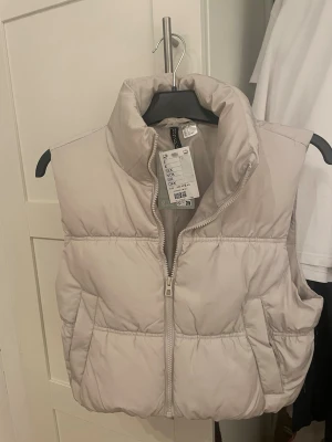 Beige dunväst från H&M - Snygg beige dunväst från H&M med hög krage och dragkedja framtill. Västen har två sidofickor 