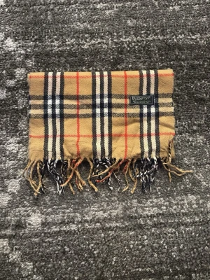 Burberrys beige rutig halsduk - Klassisk beige halsduk från Burberrys i 100% lammull med ikoniskt rutmönster i svart, vitt och rött. Halsduken har fransar i ändarna och en vävd logotypetikett. Perfekt accessoar för att lyfta din outfit.