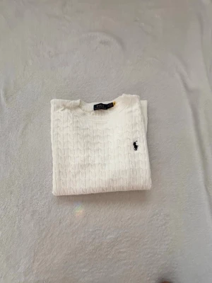 Vit kabelstickad tröja från Polo Ralph Lauren - Vit kabelstickad tröja från Polo Ralph Lauren med rund halsringning och svart broderad logga på bröstet. Tröjan har långa ärmar och ribbade muddar vid ärmslut och nederkant. Perfekt för en stilren och klassisk look. Storlek M men känns som S