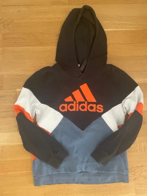 Adidas hoodie svart/blå/orange - Snygg hoodie från Adidas i svart, blått, vitt och orange. Stor Adidas-logga framtill i orange. Hoodien har huva och coola färgblock på ärmarna. Perfekt för dig som gillar sportig och avslappnad stil.