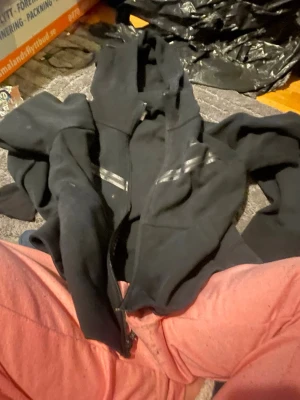 Svart hoodie med dragkedja från Nike - Säljer en svart hoodie från Nike i storlek XS. Hoodien har dragkedja framtill, två fickor och reflekterande detaljer på bröstet. Klassisk huva och mjukt material som passar perfekt till chill eller träning.