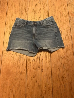Blå jeansshorts med rå kant - Snygga blå jeansshorts med rå, fransig kant och klassisk femficksdesign. Shortsen har normal passform och bälteshällor i midjan. Perfekta för varma dagar och enkel att matcha med olika toppar.
