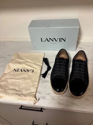 Svarta sneakers från Lanvin - Snygga svarta sneakers från Lanvin med ovandel i mocka och lackad tå. Skorna har vita sulor och klassisk snörning. Skicket är 6/10. Perfekta för dig som gillar stilrena och exklusiva detaljer. Allt og medföljer.