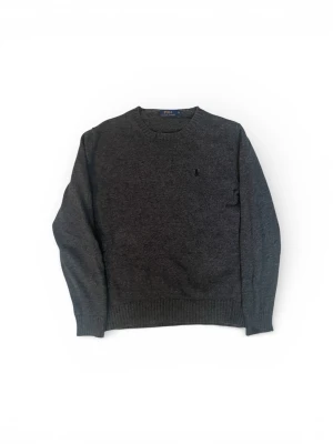 Mörkgrå tröja från Polo Ralph Lauren - Mörkgrå stickad tröja från Polo Ralph Lauren med rund halsringning och diskret broderad logga på bröstet. Tröjan har ribbade muddar vid ärmslut och nederkant, perfekt för lager-på-lager och en clean look.